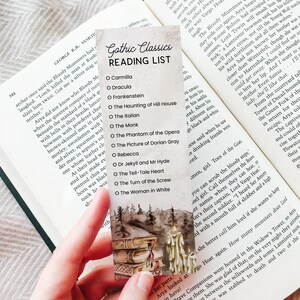 May include: A bookmark featuring a watercolour illustration of a stack of books and a candle. The bookmark has a white background with a dark brown border. The text "Gothic Classics Reading List" is printed in black at the top of the bookmark. The list of books includes "Carmilla", "Dracula", "Frankenstein", "The Haunting of Hill House", "The Italian", "The Monk", "The Phantom of the Opera", "The Picture of Dorian Gray", "Rebecca", "Dr Jekyll and Mr Hyde", "The Tell-Tale Heart", "The Turn of the Screw", and "The Woman in White".
