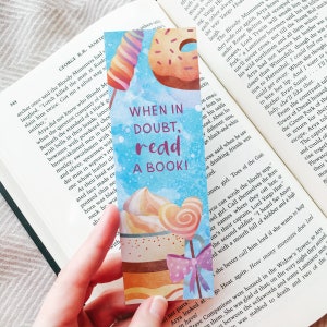 K&ouml;nnte beinhalten: Ein farbenfrohes Lesezeichen mit blauem Hintergrund und dem Text "Im Zweifel, lies ein Buch!" Das Lesezeichen zeigt Illustrationen verschiedener Desserts, darunter ein Donut, ein Cupcake und ein Lollipop.
