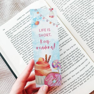 K&ouml;nnte beinhalten: Ein farbenfrohes Lesezeichen mit einem verspielten Design, das einen Donut, einen Cupcake und einen Lollipop zeigt. Das Lesezeichen tr&auml;gt den Text "Life is Short. Keep reading!" in rosa und schwarzen Buchstaben.