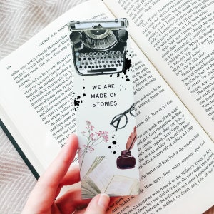 May include: A bookmark with a typewriter, inkwell, glasses, and floral illustrations. The text "WE ARE MADE OF STORIES" is displayed. The bookmark is held over an open book.