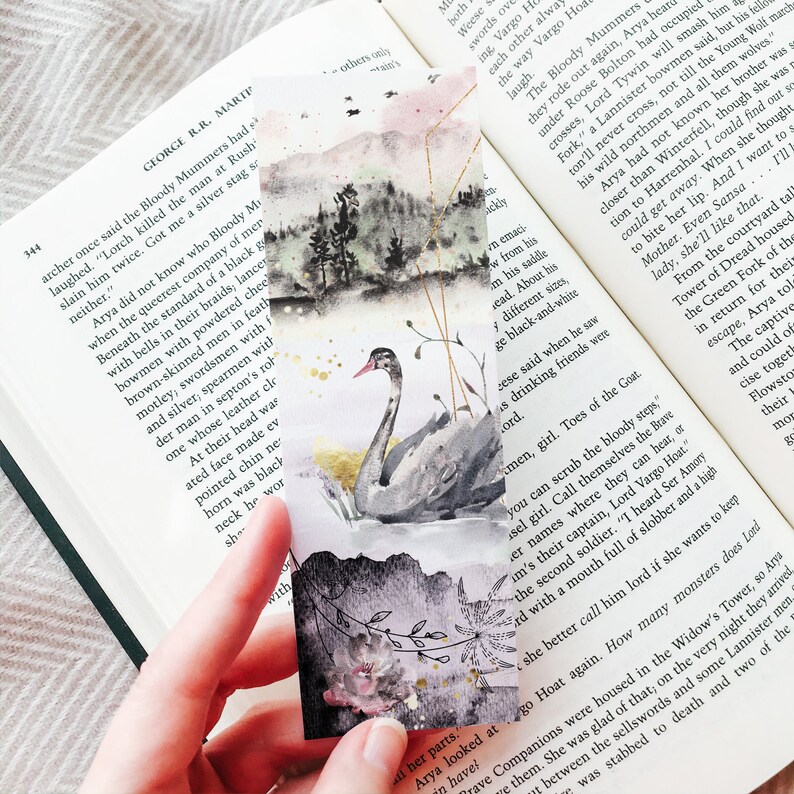 May include: A rectangular paper bookmark featuring a watercolor illustration of a black swan swimming in water. The background displays a misty mountain landscape with pine trees, gold geometric lines, and a dark floral pattern at the bottom.