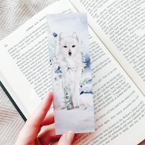 Op de afbeelding: Rechthoekige aquarel bladwijzer met een witte poolvos met bruine ogen, omringd door blauwe en groene bloemaccenten. De bladwijzer wordt in een hand gehouden, met tekst uit een boek op de achtergrond.
