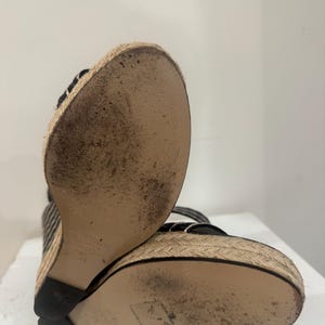 May include: Black and white striped wedge sandals with woven jute soles. The soles show wear and tear. The sandals have a black heel and straps. The shoes are angled towards the viewer.
