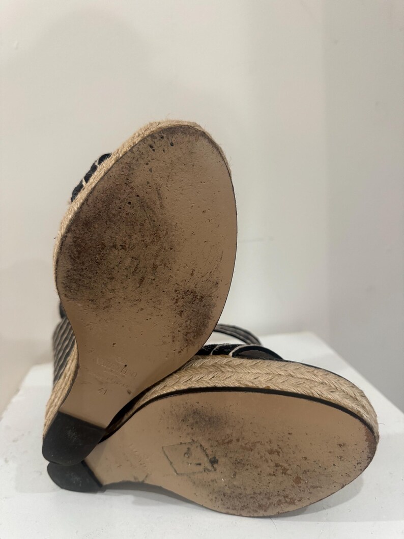 May include: Black and white striped wedge sandals with woven jute soles. The soles show wear and tear. The sandals have a black heel and straps. The shoes are angled towards the viewer.