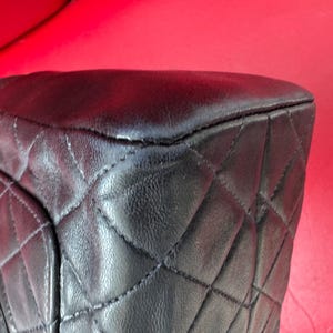 May include: Close-up of a black leather handbag with a quilted pattern. The bag's corner is visible, showing the stitching and texture of the leather. The background is a solid red.