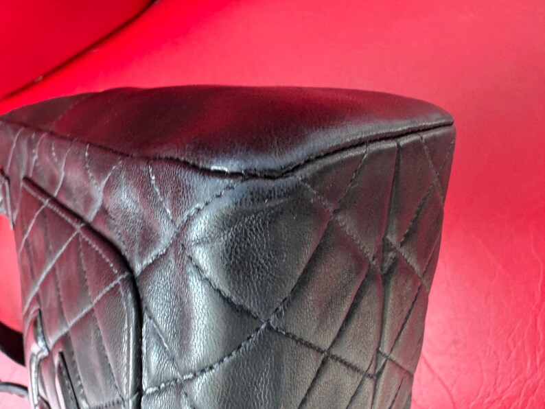May include: Close-up of a black leather handbag with a quilted pattern. The bag's corner is visible, showing the stitching and texture of the leather. The background is a solid red.