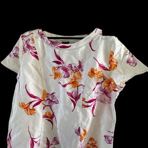 May include: A short-sleeved white blouse featuring a floral pattern with orange and magenta flowers and green stems. The garment has a round neckline with decorative stitching along the collar and vertical seams running down the front panel.