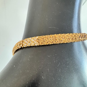 K&ouml;nnte beinhalten: Ein goldfarbenes Choker-Halsband mit strukturierter Oberfl&auml;che. Die Halskette besteht aus kleinen, eng aneinander liegenden Elementen, die ein detailliertes Muster bilden. Der Schmuck wird auf einer schwarzen Schaufensterpuppe pr&auml;sentiert.