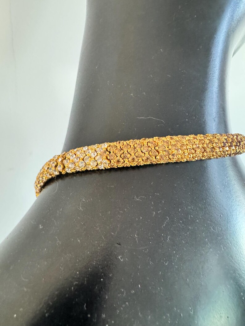 K&ouml;nnte beinhalten: Ein goldfarbenes Choker-Halsband mit strukturierter Oberfl&auml;che. Die Halskette besteht aus kleinen, eng aneinander liegenden Elementen, die ein detailliertes Muster bilden. Der Schmuck wird auf einer schwarzen Schaufensterpuppe pr&auml;sentiert.