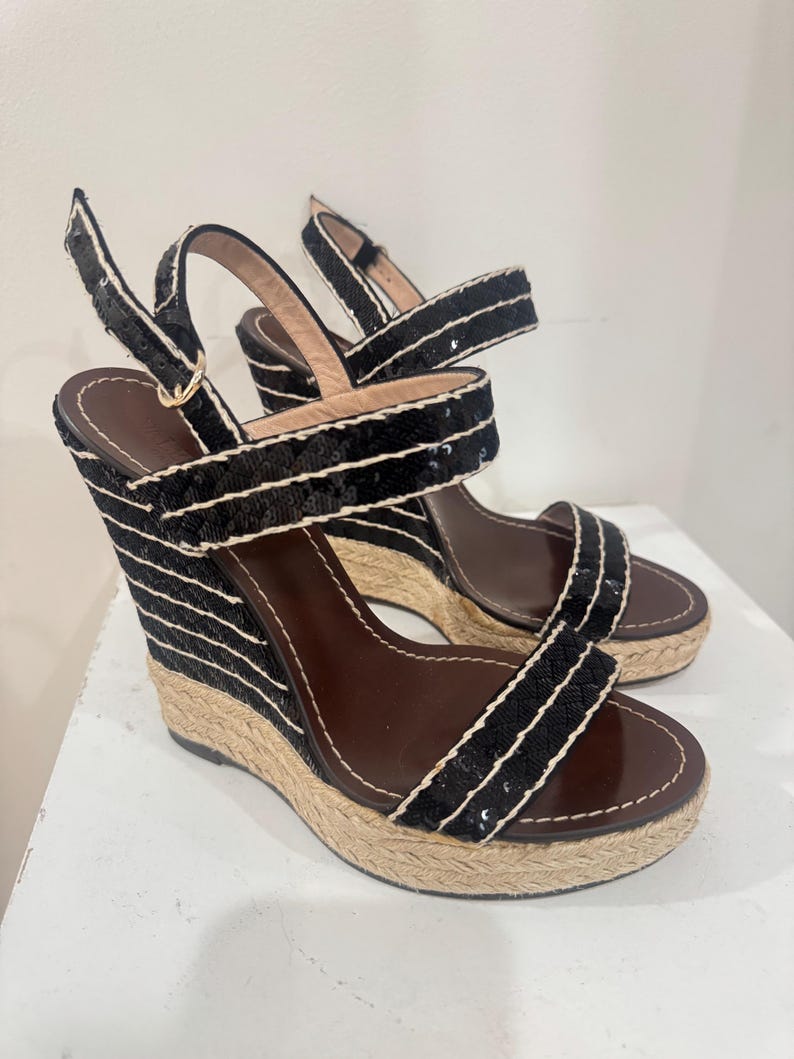 May include: Pair of black sequin wedge sandals with white trim and brown insoles. The wedge heels have a woven texture. The sandals have adjustable ankle straps and a classic design.