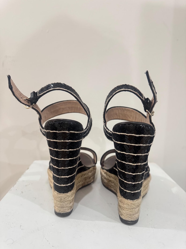May include: Pair of black sequined wedge sandals with white horizontal stripes. The sandals have ankle straps and woven jute soles. The heels are approximately 4 inches high.