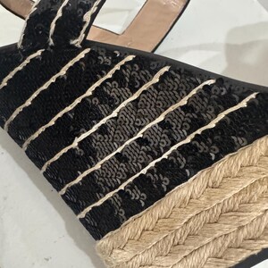May include: Black sequined wedge sandals with a woven jute sole. The sandals feature black sequins with white stitching and a tan ankle strap. The wedge heel is made of woven jute material.