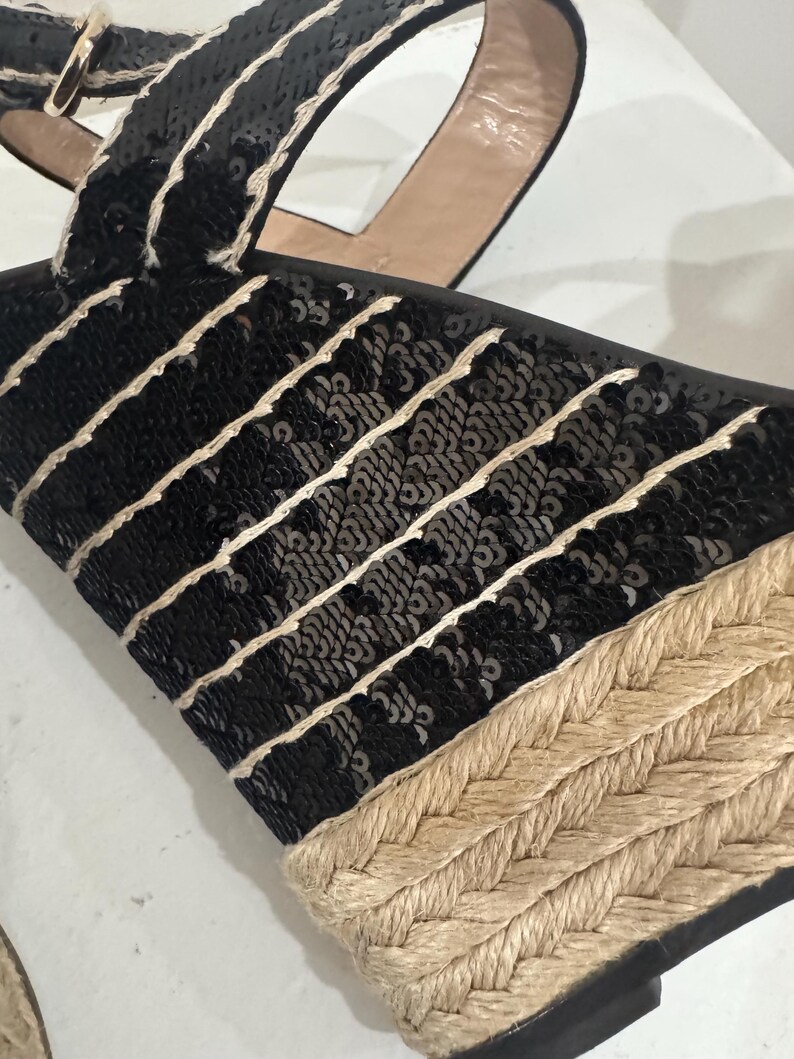 May include: Black sequined wedge sandals with a woven jute sole. The sandals feature black sequins with white stitching and a tan ankle strap. The wedge heel is made of woven jute material.