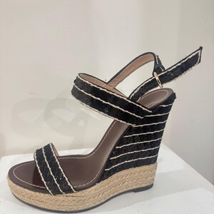 May include: Black wedge sandals with a woven jute sole and black and white striped fabric detailing. The sandals have a brown leather insole and a buckle closure ankle strap. The wedge heel provides a comfortable lift.