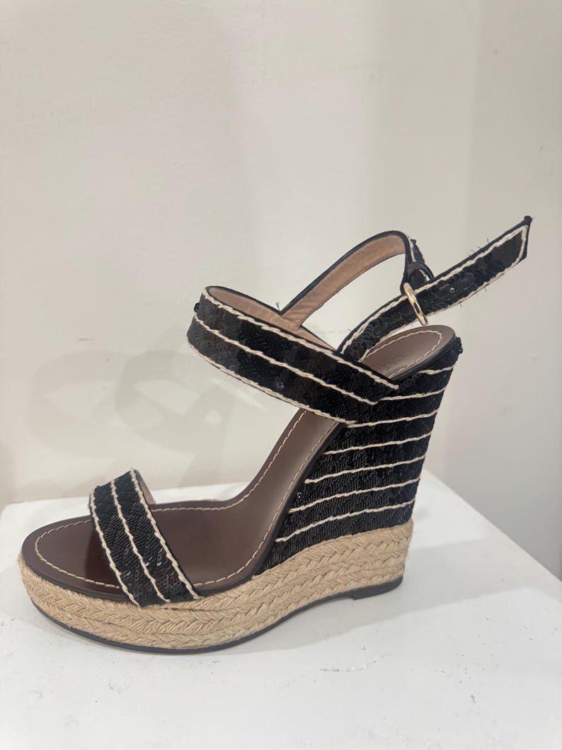 May include: Black wedge sandals with a woven jute sole and black and white striped fabric detailing. The sandals have a brown leather insole and a buckle closure ankle strap. The wedge heel provides a comfortable lift.