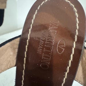 May include: Close-up of a brown leather sandal in a top-down view. The sandal features the brand name "VALENTINO GARAVANI" embossed in gold lettering. The edges are stitched with a light-colored thread, and the sandal has a dark brown sole.