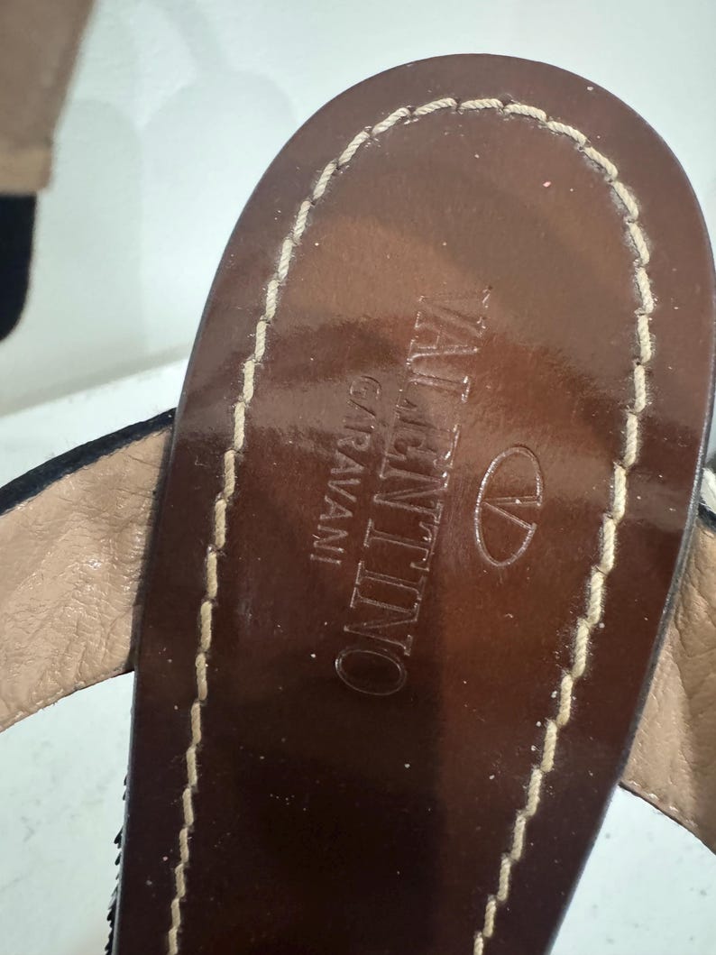 May include: Close-up of a brown leather sandal in a top-down view. The sandal features the brand name "VALENTINO GARAVANI" embossed in gold lettering. The edges are stitched with a light-colored thread, and the sandal has a dark brown sole.
