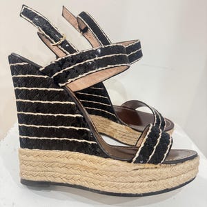 May include: Black wedge sandals with sequin and rope detailing. The sandals feature a platform sole with a woven texture. The straps are black with white accents and a buckle closure. The wedge heel is approximately 4 inches.
