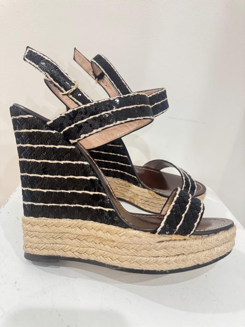May include: Black wedge sandals with sequin and rope detailing. The sandals feature a platform sole with a woven texture. The straps are black with white accents and a buckle closure. The wedge heel is approximately 4 inches.