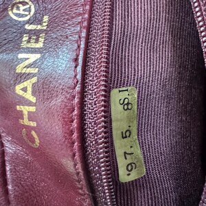 May include: Close-up of a burgundy leather interior of a handbag. The CHANEL logo is embossed in gold. A small tag with the numbers "97. 5. 8. I" is visible. The interior features a zipper and a textured fabric lining.