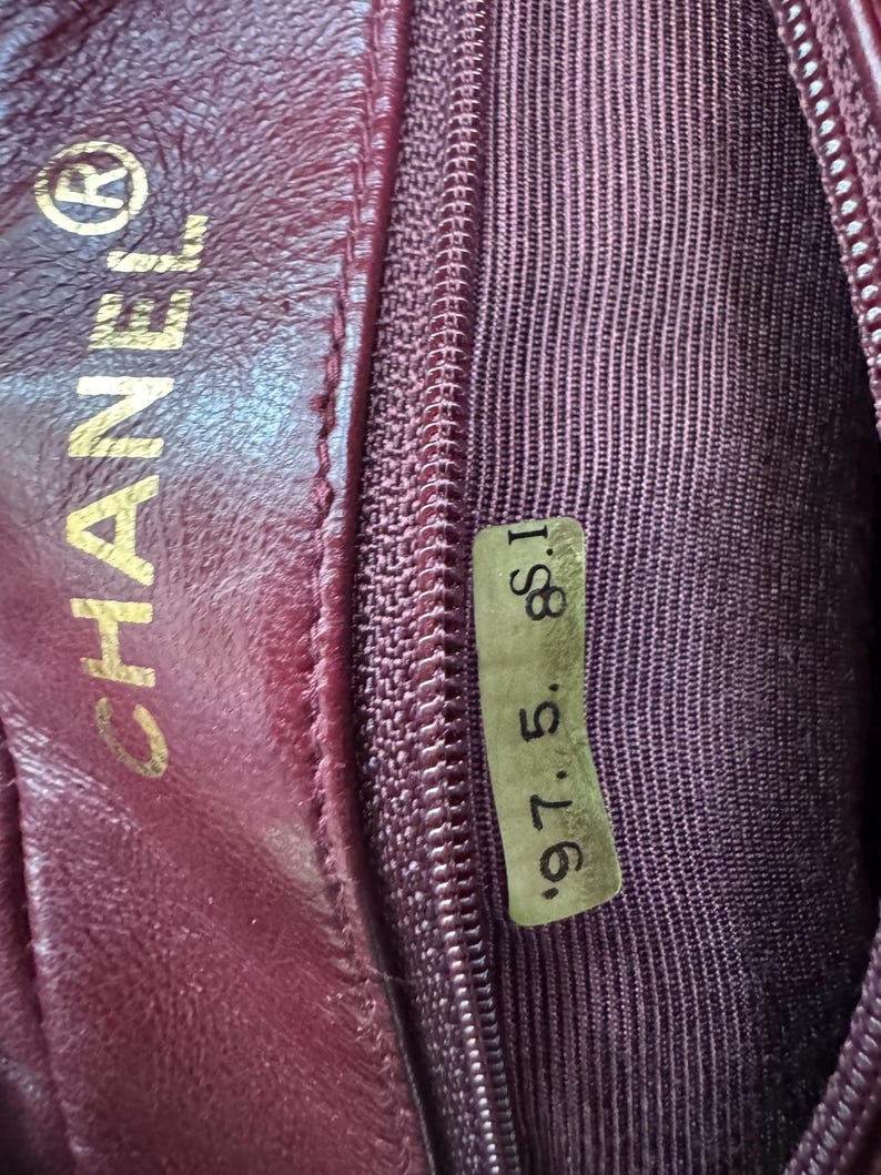 May include: Close-up of a burgundy leather interior of a handbag. The CHANEL logo is embossed in gold. A small tag with the numbers "97. 5. 8. I" is visible. The interior features a zipper and a textured fabric lining.