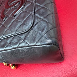 May include: Black quilted leather handbag with a flap closure and a large, glossy logo. The bag features a diamond pattern and gold-toned hardware at the base. The bag is set against a red background.