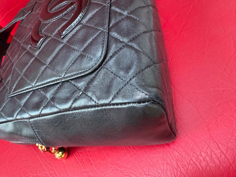 May include: Black quilted leather handbag with a flap closure and a large, glossy logo. The bag features a diamond pattern and gold-toned hardware at the base. The bag is set against a red background.