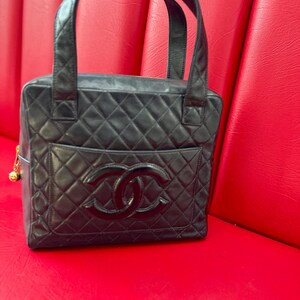 May include: Black quilted leather handbag with top handles. The bag features a diamond pattern, a front pocket, and a large interlocking logo. A gold-colored charm hangs from the side. The bag is sitting on a red surface.