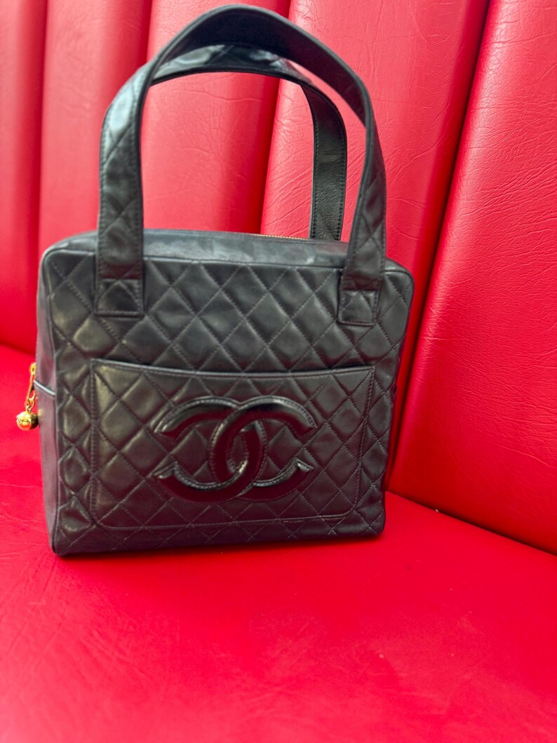May include: Black quilted leather handbag with top handles. The bag features a diamond pattern, a front pocket, and a large interlocking logo. A gold-colored charm hangs from the side. The bag is sitting on a red surface.