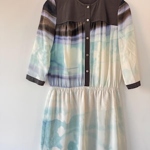 May include: A button-down dress with a watercolor-style print in shades of blue, green, and brown. The dress has a dark brown yoke and sleeves with a gathered waist. The dress is hanging on a wooden hanger.