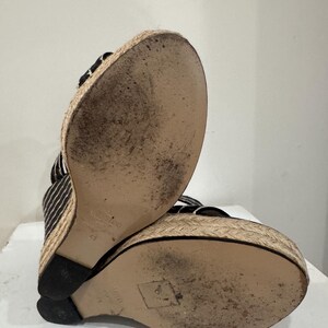 May include: Black and white striped wedge sandals with a woven jute platform and sole. The sandals have a black heel and straps. The soles show signs of wear.