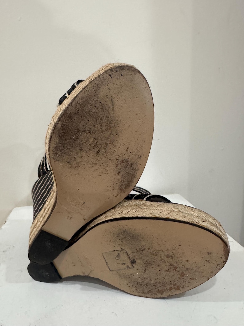 May include: Black and white striped wedge sandals with a woven jute platform and sole. The sandals have a black heel and straps. The soles show signs of wear.