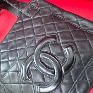 May include: Black quilted leather tote bag with a large interlocking logo. The bag features a diamond pattern and a top handle. The bag is on a red surface.