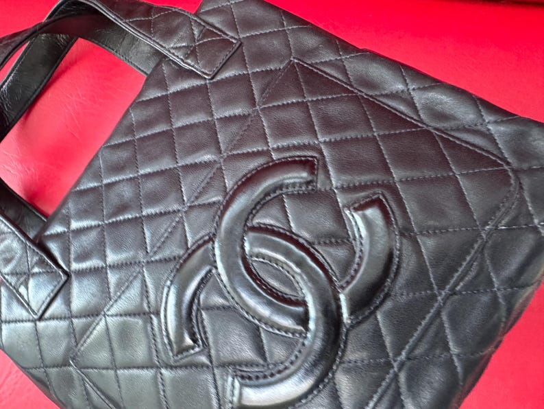 May include: Black quilted leather tote bag with a large interlocking logo. The bag features a diamond pattern and a top handle. The bag is on a red surface.
