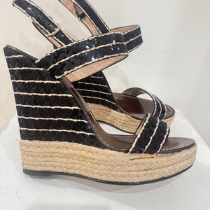 May include: Black wedge sandals with a woven jute sole and black sequin straps. The sandals have a buckle closure and a brown insole. The wedge heel is approximately 4 inches high.