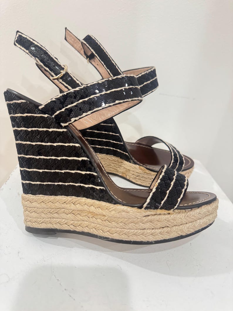 May include: Black wedge sandals with a woven jute sole and black sequin straps. The sandals have a buckle closure and a brown insole. The wedge heel is approximately 4 inches high.