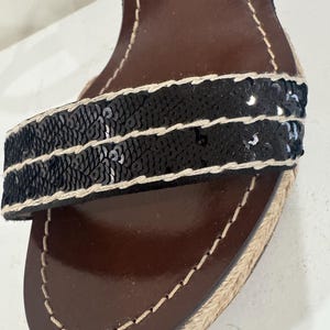 May include: Close-up of a black sequined sandal strap with a brown leather insole. The strap is embellished with rows of black sequins and bordered with beige stitching. The sole is trimmed with woven jute.