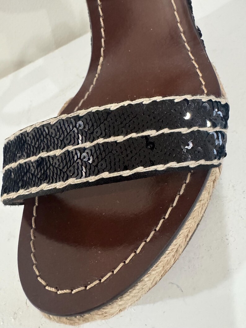 May include: Close-up of a black sequined sandal strap with a brown leather insole. The strap is embellished with rows of black sequins and bordered with beige stitching. The sole is trimmed with woven jute.