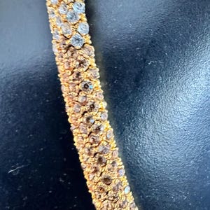 K&ouml;nnte beinhalten: Nahaufnahme eines goldfarbenen Armbands mit Reihen aus funkelnden, klaren und braunen Kristallen. Die Kristalle sind dicht gesetzt und bilden eine durchgehende, schimmernde Oberfl&auml;che. Das Armband befindet sich auf dunklem Hintergrund.