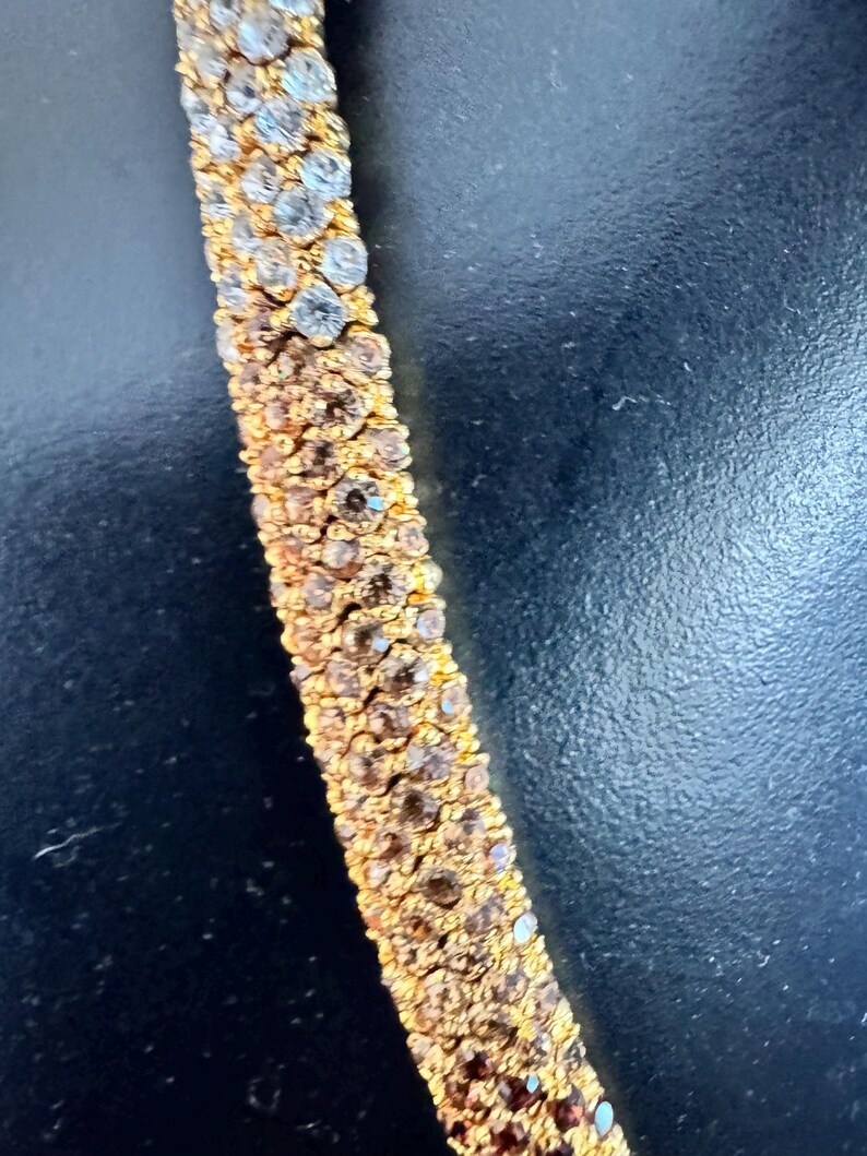 K&ouml;nnte beinhalten: Nahaufnahme eines goldfarbenen Armbands mit Reihen aus funkelnden, klaren und braunen Kristallen. Die Kristalle sind dicht gesetzt und bilden eine durchgehende, schimmernde Oberfl&auml;che. Das Armband befindet sich auf dunklem Hintergrund.