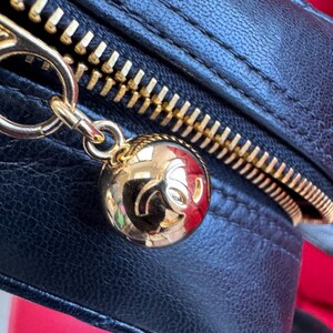 May include: Close-up of a black leather handbag with a gold zipper and a gold charm. The charm is a round, gold ball with a logo. The zipper is gold and the leather is textured.