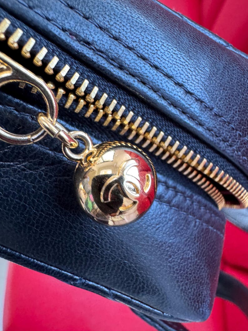 May include: Close-up of a black leather handbag with a gold zipper and a gold charm. The charm is a round, gold ball with a logo. The zipper is gold and the leather is textured.