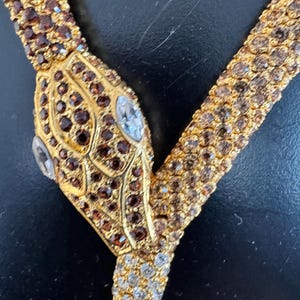 K&ouml;nnte beinhalten: Eine goldfarbene Schlangenhalskette mit braunen und klaren Kristallakzenten. Der Schlangenkopf hat braune Kristalle und zwei klare, oval geformte Kristalle als Augen. Die Halskette hat ein Y-f&ouml;rmiges Design.