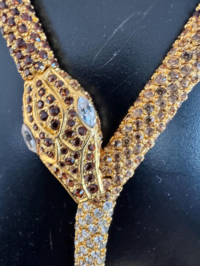 K&ouml;nnte beinhalten: Eine goldfarbene Schlangenhalskette mit braunen und klaren Kristallakzenten. Der Schlangenkopf hat braune Kristalle und zwei klare, oval geformte Kristalle als Augen. Die Halskette hat ein Y-f&ouml;rmiges Design.