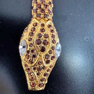K&ouml;nnte beinhalten: Ein goldfarbener Schlangenanh&auml;nger mit braunen und klaren Kristallakzenten. Der Schlangenkopf ist mit braunen Kristallen und klaren Kristallaugen versehen. Der Anh&auml;nger hat ein strukturiertes, verziertes Design.