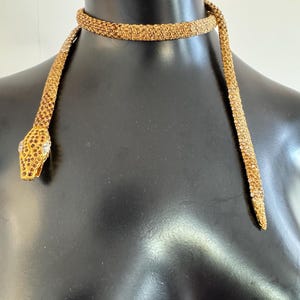 K&ouml;nnte beinhalten: Eine goldfarbene Schlangenhalskette mit detailliertem Design. Der Schlangenkopf hat ein juwelenbesetztes Auge und ein strukturiertes Muster. Die Halskette ist um eine schwarze Schaufensterpuppe drapiert.