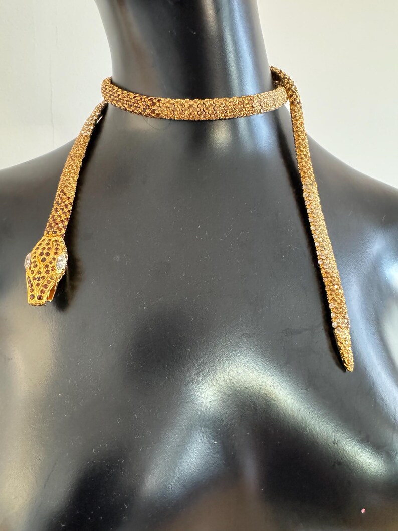 K&ouml;nnte beinhalten: Eine goldfarbene Schlangenhalskette mit detailliertem Design. Der Schlangenkopf hat ein juwelenbesetztes Auge und ein strukturiertes Muster. Die Halskette ist um eine schwarze Schaufensterpuppe drapiert.