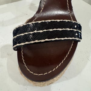 May include: Close-up of a brown leather sandal with a black sequined strap. The sandal has a woven jute sole and white stitching details. The heel is visible in the background.