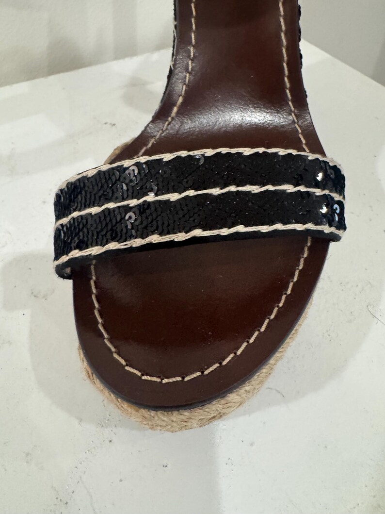 May include: Close-up of a brown leather sandal with a black sequined strap. The sandal has a woven jute sole and white stitching details. The heel is visible in the background.
