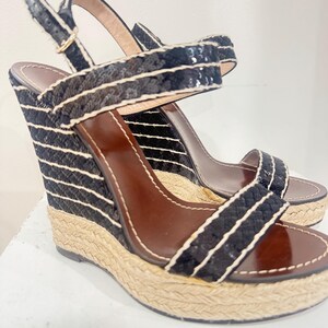 May include: A pair of black wedge sandals with white striped detailing. The sandals have a woven jute platform and a brown leather insole. The straps are black with sequin accents and a gold buckle.